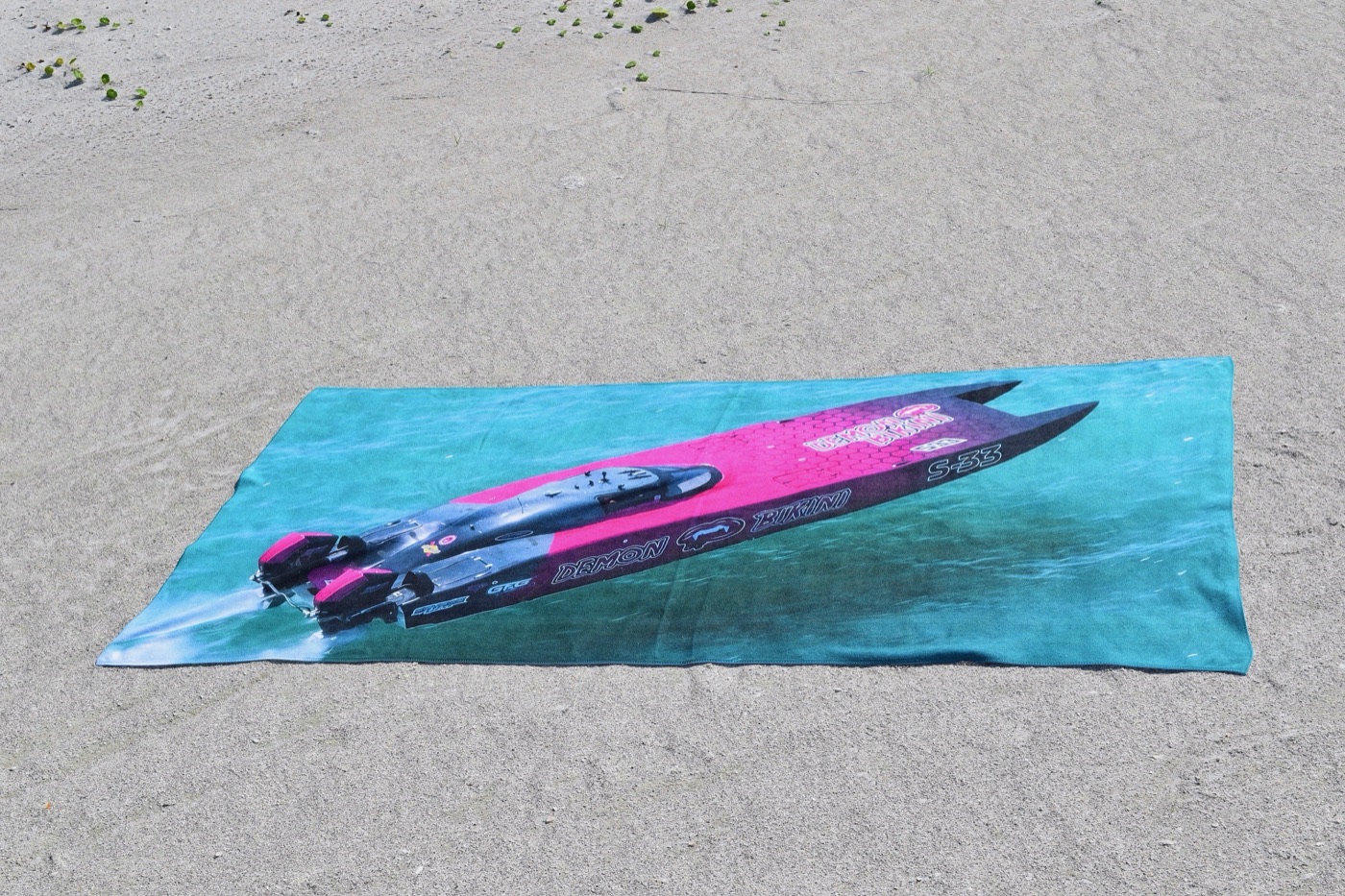 Beach towel printed with the pink and black Demon Bikini S-33 race boat on turquoise water, laid out on sand